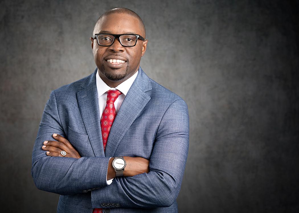 Dr. Jermaine Whirl, a smiling African American male, wearing a light blue suit jacket, white collared shirt and red tie with a white flowered print smiles at the camera pictured against a grey background.